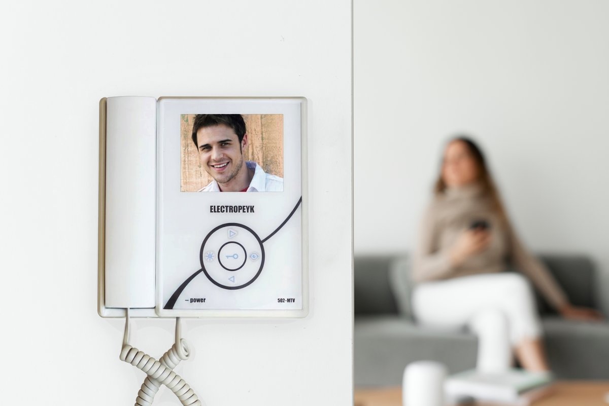 Black video door phone unit installed on a wall, with a person answering in the background