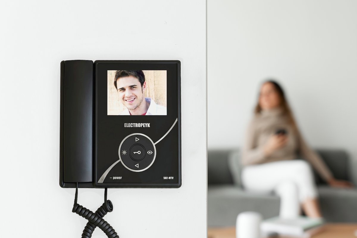 White video door phone unit installed on a wall, with a person answering in the background
